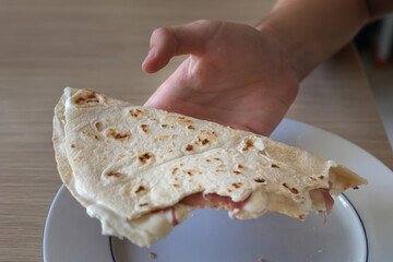 Hand Holding a Bitten Flat Bread Filled with Cheese and Ham on top of a Plate