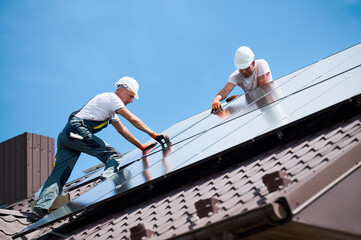 Workers building solar panel system on rooftop of house. Two men installers in helmets installing photovoltaic solar module outdoors. Alternative, green and renewable energy generation concept.