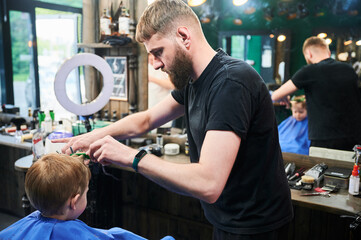 Professional hairdresser making haircut for his little client. Male barber using hairdresser's tools. Back view of little boy getting his first haircut.