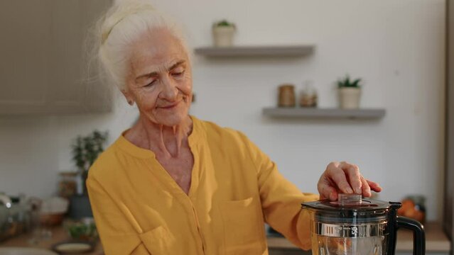Tilting up medium shot of happy Caucasian elderly woman making healthy smoothie in blender at modern kitchen
