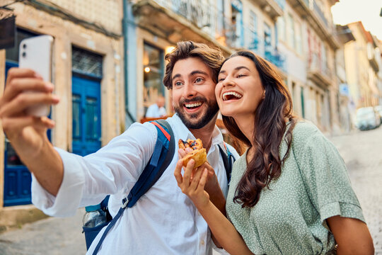 Cheerful couple holding traditional dessert pastel de nata and taking selfie through smart phone