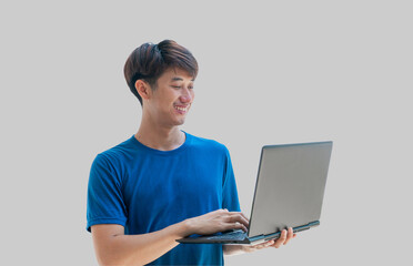 A young Asian man in his 20s wearing a blue t-shirt using a laptop for online remote work isolated on a gray background. Man watching webinars on a laptop, e-learning, e-banking.