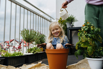 Father watering playful girl sitting in pot at balcony