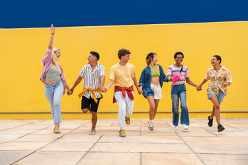 Multi-ethnic group of young friend running in front of yellow wall holding hands