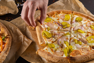 a man's hand pulls a piece of crispy baked toasted pizza with ham, pickled cucumbers and onions. on a dark background