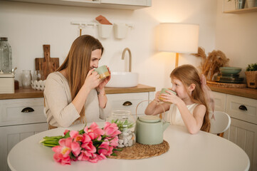 Obraz na płótnie Canvas Happy mother with daughter drink tea or cocoa sitting in kitchen and having fun. Family communication. Mom and children spend time together. Child congratulates mother with flowers on Mother's Day.