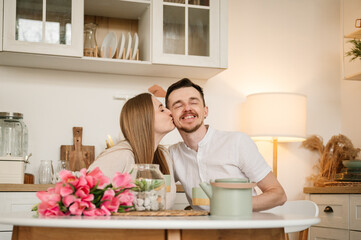 Obraz na płótnie Canvas Romantic female kisses male, spending time together. Loving young couple having conversation and drink tea in morning. Couple drinking coffee in the kitchen.