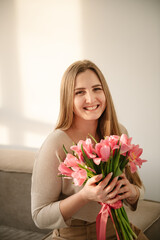 Portrait of a young girl in dress holding hands bouquet of pink tulips. Beautiful woman is smelling a big bunch of spring flowers and sitting on sofa at home. International Women's Day. Looking camera