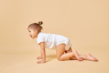 Side view portrait of pretty little baby, girl crawling across floor in cute white romper against...