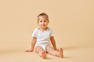 Portrait of joyful little baby in white romper sitting on floor with cute cheerful smile against beige studio background. Concept of childhood, motherhood, life, birth. Copy space for ad
