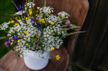 White vase with meadow flowers, herbs and grass, placed on a wooden chair. A colorful variety of summer wildflowers.
