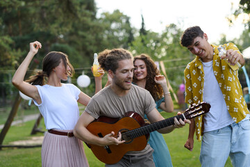 Handsome man playing at guitar for dancing friends at party. Friends and family dancing and having fun at summer grill garden party.