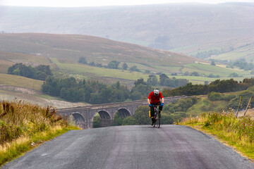 Tired cyclist pedals on at the top of a very steep hill in the Yorkshire Dales National Park, with Dent Head viaduct in the background