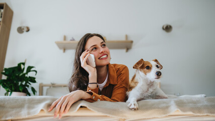 Beautiful single woman lying on bed, petting her dog, and making phone call. Young woman living...