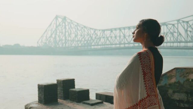 A young attractive Indian traditional Hindu woman in an ethnic saree standing outside on the riverbank enjoying the view of the Ganges or Hooghly River and Howrah Bridge is visible in the background.