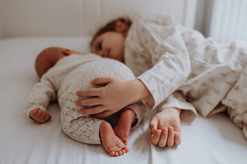 Portrait of big sister cuddling newborn, little baby. Girl lying with her new sibling in bed, closed eyes. Sisterly love, joy for new family member.