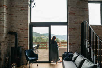 Young woman staying on terrace of wooden modern house with panoramic windows near pine forest. Concept of solitude and recreation on nature. Wellness and mindful resort. Beautiful place for vacation.