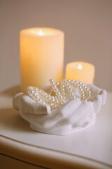 Pearl necklace lying in the plaster vase in the shape of hands on burning candles background. Beaty...