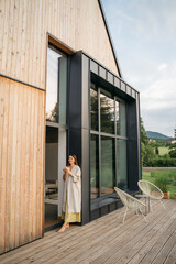 Young woman staying on terrace of wooden modern house with panoramic windows near pine forest. Concept of solitude and recreation on nature. Wellness and mindful resort. Beautiful place for vacation.