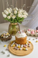 Obraz na płótnie Canvas Easter composition on the dining table. Easter cake decorated with colored pastel eggs on a wooden tray. Spring flowers, tulips and willow branches, Easter decor, composition, painted eggs. Soft focus