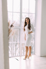 young long-haired girl in a white dress and high-heeled shoes stands in a bright room against the background of a window