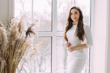 young long-haired girl in a white dress stands in a bright room against the background of a window