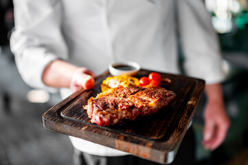A person in a white outfit holds a wooden tray with a grilled steak, sauce, and vegetables. The background is blurred, suggesting an indoor setting, possibly a restaurant or bar - Powered by Adobe