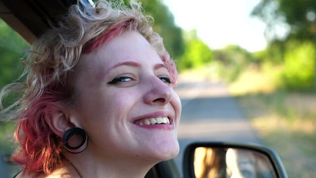 Close up to head of happy punk girl with ear tunnels leaning out of auto window and looking into camera. Smiling hippie woman enjoying car journey at summer day. Adventure and travel concept. Slow mo