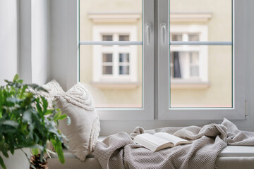 Windowsill with decorative cushions, soft plaid and open book in cozy apartment