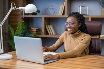 Cheerful woman freelancer in glasses with dreadlocks sitting at workplace with laptop