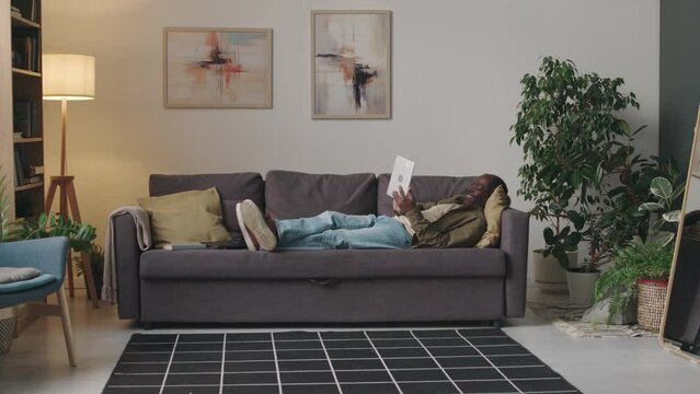 Full stab shot of adult Black man lying relaxedly on couch in modern apartment at daytime and video calling on digital tablet computer