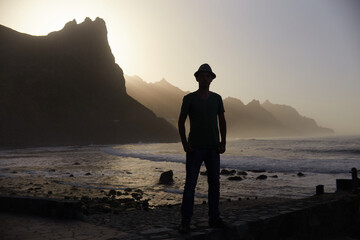 Silhouette of a man with a hat stands on a rocky coast, with mountains and a dusk sky as a backdrop