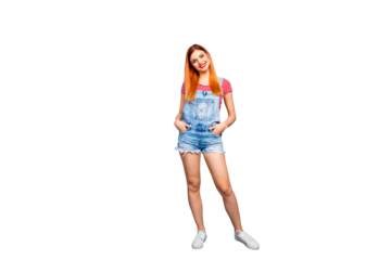 Full-body portrait of charming young girl stands isolated on bright blue background with hands in pockets of blue overalls