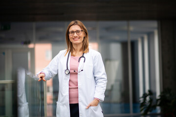 Portrait of female doctor standing in hospital, leaning on glass railing, looking at camera.