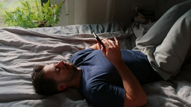 Bored man lying down on bed and browsing mobile phone