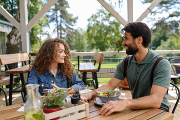 Young couple laughing at date in restaurant, sitting on restaurant terrace. Boyfriend and girlfriend enjoying springtime, having lunch outdoors.