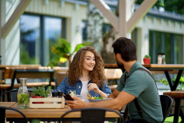 Young couple laughing at date in restaurant, sitting on restaurant terrace. Boyfriend and girlfriend enjoying springtime, having lunch outdoors.