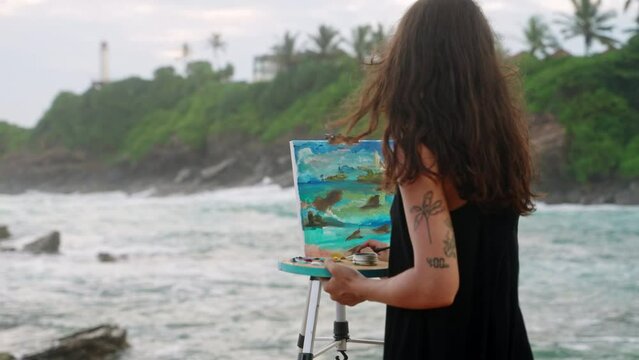 Artist paints vibrant seascape on canvas at rocky coastline. Waves crash, inspire female painter outdoor art session. Hobbyist creates impressive ocean scene near lighthouse, reflecting stormy mood.