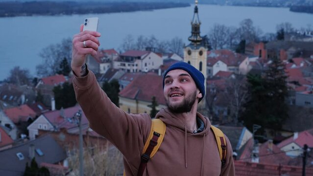 Young tourist man taking selfie using smartphone in old town Zemun in Belgrade, Serbia. Front view. Red tiled roofs of houses, top view from observation deck. Church and Danube river behind