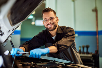 A portrait of a mechanic man smiling for the camera while working on a car, using some tools. - 761358313
