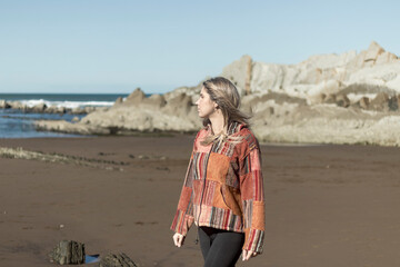 A woman wearing a patchwork jacket is walking on a beach. The beach is rocky and the sky is clear