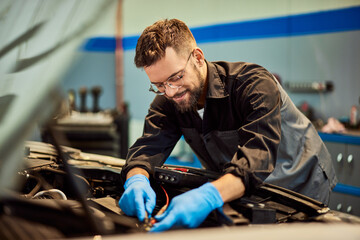 A smiling mechanic fixing a car, holding some cables in the opened hood. - 761358769