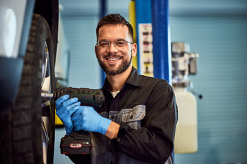 Portrait of a smiling mechanic, using a drill, screwing the wheel, and working in his garage. - 761358911