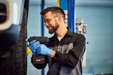 A smiling mechanic man using a drill, screwing the wheel. - 761358942
