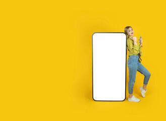 Digital mobile application advertisement concept idea image. Full body young woman 20s leaning big empty screen cell phone direct thumb display touchscreen mock up. Yellow studio background copy space
