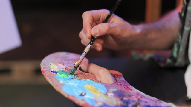 Close up view of artist hands holding paintbrush and mixing paints on colorful palette in studio. Skilled caucasian male preparing to creating masterpiece indoors. Creative space.