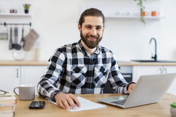 Fototapeta premium Efficient business owner improving contract while checking data on portable computer.Handsome Caucasian man wearing checkered shirt writing on document while staying at home during midday.