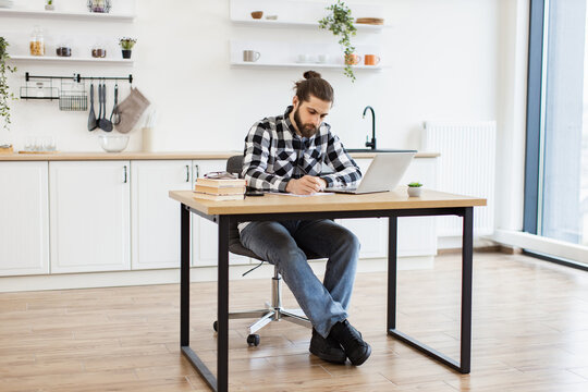 Full length of attractive Caucasian businessman sitting at office desk with digital devices in spacious dining room. Self-employed worker carrying out professional activity in comfortable workplace.