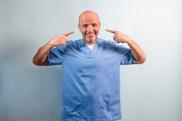 Portrait of a physiotherapist wearing light blue gown and pointing at his face with his fingers.