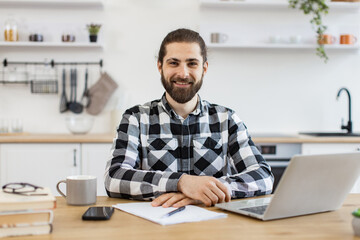 Fototapeta premium Confident Caucasian man in checkered shirt using laptop while sitting at desk on kitchen background, looking at camera. Efficient freelancer searching information while doing full-time job in home.